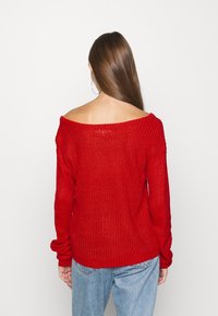 Roter Strickpullover mit Carmen-Ausschnitt, langen Ärmeln, gerippter Textur und lockerer Passform, kombiniert mit hellblauen Jeans.