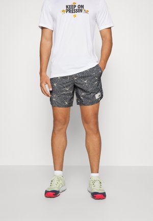 Harmaat shortsit, joissa on valkoinen abstrakti kuvio, yhdistettynä valkoiseen t-paitaan, jossa on värikäs iskulause. Jalkineet ovat vaaleanvihreät, joissa on punaisia yksityiskohtia.