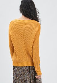 Senfgelbes Strickpullover mit weit geschnittenem Ausschnitt, gerippten Bündchen und einem strukturierten Muster. Getragen über einem Rock mit Blumenmuster.
