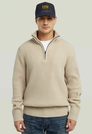 Mann in einem beigen, gerippten Half-Zip-Pullover, blauen Jeans und navyblauer G-Star Originals Kappe, stehend vor einem schlichten hellen Hintergrund.