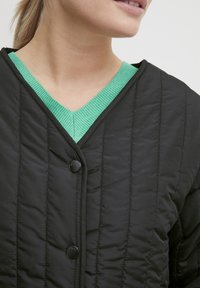 Laget svart jakke med V-hals, grønn ribbestrikket krage og runde svarte knapper, som viser tekstur og en tettsittende design.