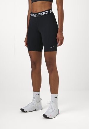 Sportler trägt schwarze Nike Pro Shorts, weiße Nike-Socken und weiße Sportschuhe, steht auf einem weißen Boden vor einem weißen Hintergrund.