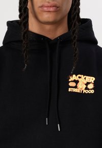 Svart huvtröja med grafiskt tryck i orange accenter, som visar texten "Jacker Street Food" och en hund som bär kockmössa.