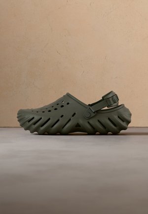Chaussure à enfiler vert foncé avec des trous de ventilation, une semelle texturée et une bride arrière réglable sur une surface et un fond neutres.