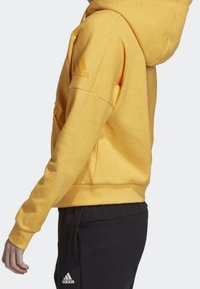 Žlutá mikina s kapucí a strukturovaným povrchem, s kapsou typu klokanka a žebrovanými manžetami. Černé tepláky s viditelným logem.