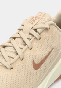 Chaussure de sport beige avec un dessus en maille texturée, des lacets ton sur ton et un swoosh Nike marron. Semelle rembourrée et design minimaliste.