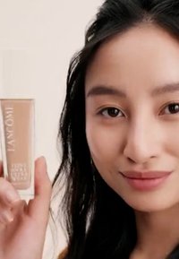 Bouteille de fondation en verre transparent avec un liquide beige clair, de forme rectangulaire, étiquette indiquant "LANCOME," tenue par une main contre un fond neutre.