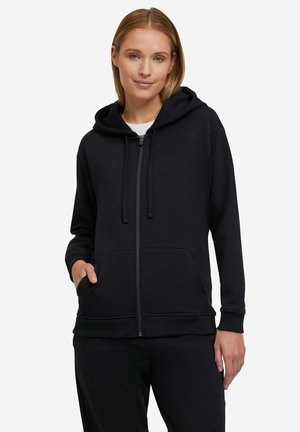 Vrouw met een zwart hoodie met ritssluiting en zakken aan de voorkant, gecombineerd met zwarte broek, staand tegen een witte achtergrond.