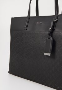 Calvin Klein svart tote bag laget av teksturert syntetisk materiale, med en glatt topp, preget logo og en avtakbar etikett.