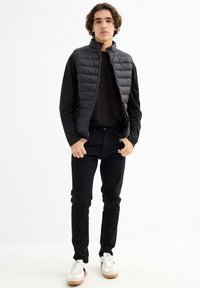 Chaleco acolchado negro con diseño de líneas horizontales, llevado sobre una camiseta de manga larga negra, combinado con pantalones negros y zapatillas blancas.