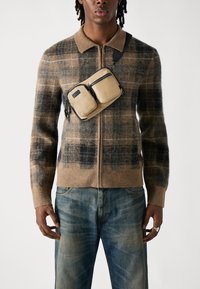 Cardigan a zip con motivo a quadri marroni realizzato in morbida lana, abbinato a jeans blu sbiaditi. Una borsa a tracolla marrone chiaro con due tasche con zip è indossata sul petto.