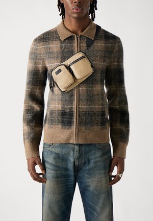 Cardigan a zip con motivo a quadri marroni realizzato in morbida lana, abbinato a jeans blu sbiaditi. Una borsa a tracolla marrone chiaro con due tasche con zip è indossata sul petto.