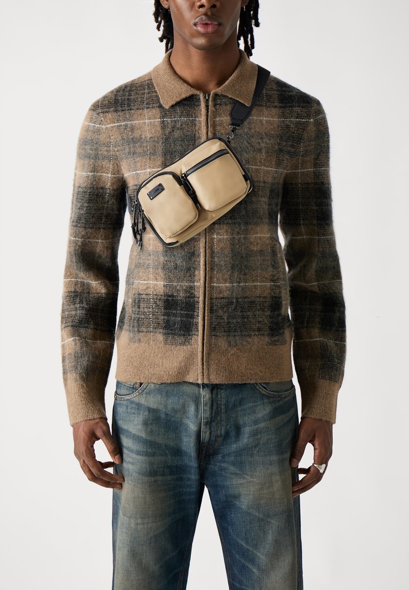 Cardigan a zip con motivo a quadri marroni realizzato in morbida lana, abbinato a jeans blu sbiaditi. Una borsa a tracolla marrone chiaro con due tasche con zip è indossata sul petto.