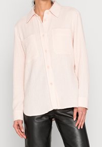 Mujer que lleva una camisa de botón de manga larga texturizada en rosa claro con dos bolsillos en el pecho y pantalones de cuero negros, de pie contra un fondo blanco.