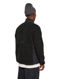 Mand iført sort fleecejakke, grå hue og sorte bukser, vendt væk fra kameraet mod en hvid baggrund.