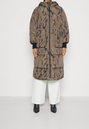 Long manteau matelassé marron et noir à motifs, avec capuche, fermeture éclair devant, grandes poches à rabat, porté avec un pantalon blanc à jambes larges et des bottes pointues noires.