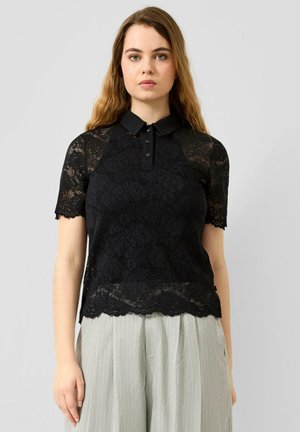 Junge Frau mit langen braunen Haaren, die ein schwarzes Spitzen-Poloshirt mit kurzen Ärmeln und hellgraue plissierte Hose trägt, steht vor einem grauen Hintergrund.