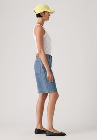 Νεαρή γυναίκα που φοράει κίτρινο καπέλο, λευκό αμάνικο τοπ, μπλε τζιν φούστα μέχρι τα γόνατα και μαύρα φλατ παπούτσια, στέκεται προφίλ σε απλό φόντο.