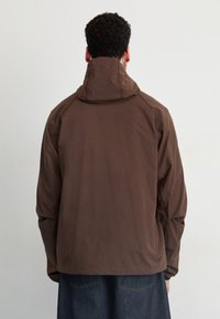 Brown lightweight jacket with a high collar and hood. Features raglan sleeves and a smooth texture. Logo detail near the hem.