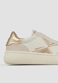 Sneaker beige con tomaia in pelle liscia, dettagli dorati sul tallone e sul logo, e suola bianca spessa e testurizzata.