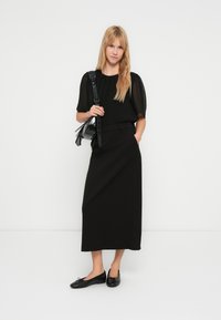 Schwarzes Kurzarm-Top mit transparenten Ärmeln, kombiniert mit einem langen schwarzen Rock und schwarzen Ballerinas. Accessoires umfassen eine kleine schwarze Handtasche.