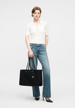 Sieviete baltā īsroku krekliņā un zilās džinsās, turot rokā lielu melnu rokas somu, stāvot pret vienveidīgu gaišu fonu.