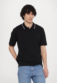 Tricou polo negru din bumbac cu o textură subtilă. Acesta dispune de un guler cu trei butoane și accente albe cu dungi pe guler.