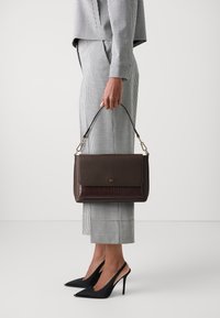 Brązowa skórzana torebka z akcentami w wzór krokodyla, wyposażona w klapę zapięcia i krótki uchwyt. Noszona z czarno-białym garniturem w kratę.