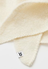 Crèmekleurige gebreide sjaal met een zachte textuur, voorzien van een gekartelde rand en een klein geweven label met een logo.