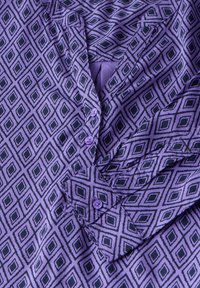 Camicia viola con un motivo a rombi in verde scuro. Presenta un colletto, una pattina con bottoni e una texture di tessuto liscia. Decorata con bottoni viola.