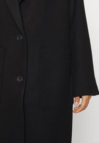 Manteau en laine noire avec une coupe décontractée, doté d'un col à revers, de deux poches avant et d'une fermeture à boutons. Tissu texturé pour plus de chaleur.