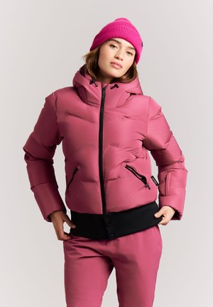 Vrouw draagt felroze muts, roze donsjas met zwarte rits en bijpassende broek, staand tegen een effen lichte achtergrond.