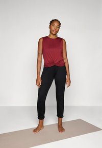 Bordeaux mouwloze top met een geknoopt detail in de taille, gecombineerd met zwarte taps toelopende broek. Gladde stof met een ontspannen pasvorm, ideaal voor sportkleding.