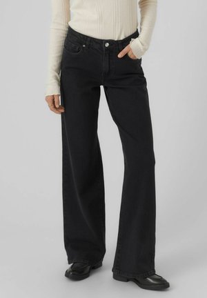 Osoba nosząca czarne jeansy o szerokich nogawkach, kremowy prążkowany top z długim rękawem, czarne buty, z jedną ręką w kieszeni i pierścionkami na palcach.