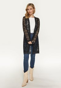 Cardigan in pizzo nero con apertura frontale, motivi dettagliati, orlo ad arco, abbinato a una maglietta bianca e jeans blu, completato da stivali beige.