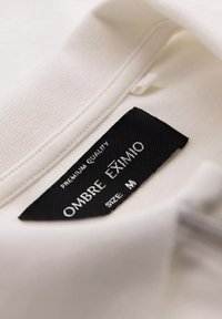 Hvidt stof med en glat tekstur, der har et sort mærke. Mærket viser "OMBRE EXIMIO" og "STØRRELSE: M" i sølvfarvet skrift.