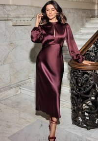 Bordo satino suknelė su aukštu iškirpimu, raktų angos detalėmis ir ilgomis pūstomis rankovėmis. Prigludusi juosmens dalis ir plazdanti sijonas, derinama su raudonais sandalais ant aukšto kulno.