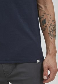 Mørkeblå bomulls-T-skjorte med rund hals og korte ermer. Har en liten etikett i fallen og en jevn tekstur. Grå bukser synlig under.
