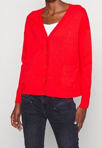Czerwony dziany kardigan z dekoltem w serek, długimi rękawami oraz kieszeniami z przodu; posiada kontrastujące guziki i luźny krój.