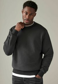 Sudadera negra con cuello redondo y puños acanalados. Lleva puesta una capa blanca, presenta una textura suave y un corte relajado.