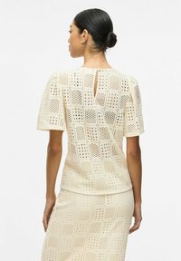 Blusa de encaje en crema claro con patrones cuadrados y detalles cortados, con mangas cortas y una apertura en forma de llave en la espalda. Textura suave.