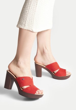 Pies con sandalias de tacón abiertas de dedo en rojo con correas anchas, acompañados de una falda blanca texturizada, sobre un fondo liso.