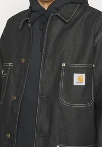 Chaqueta de denim negro con un diseño abotonado, costuras blancas y un bolsillo en el pecho izquierdo con un parche del logo de Carhartt. Sudadera con capucha debajo.