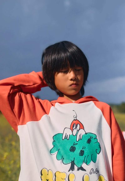 Sudadera con mangas de color coral y cuerpo blanco que muestra un personaje de dibujos animados en un árbol verde. Tejido suave con un diseño gráfico colorido.