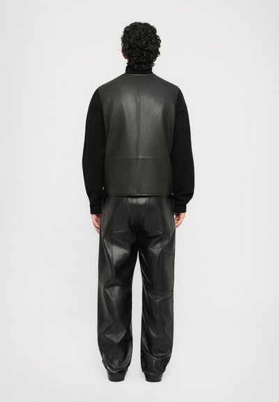 Person with curly hair wearing a black long-sleeve shirt, black leather vest, and black leather pants, standing facing away against a white background.