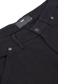 Κοντινή λήψη μαύρου παντελονιού που δείχνει την μέση, τα θηλάκια της ζώνης, ένα μεταλλικό κουμπί με λογότυπο και μια ετικέτα με την επωνυμία "FORMER".