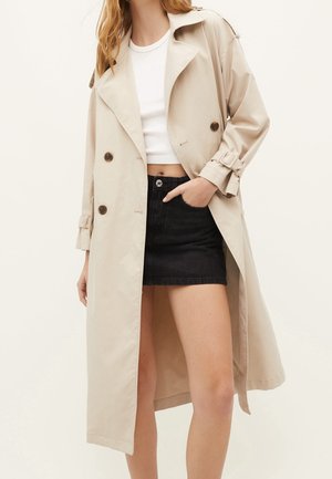 Beżowy płaszcz trencz o dwurzędowym kroju, z klapowanym kołnierzem i podwiniętymi rękawami, zestawiony z białym krótki topem i czarną spódnicą.