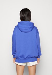 Överdimensionerad blå hoodie med en stor, mjuk huva, sänkta axlar och muddade ärmslut. Tyget verkar mjukt och strukturerat.