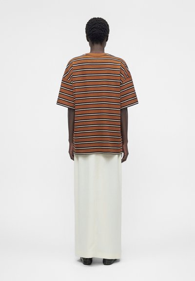 Άτομο με κοντά σγουρά μαλλιά που φοράει μια καφέ, μαύρη και λευκή ριγέ φαρδιά μπλούζα, μακριά λευκή φούστα και μαύρα παπούτσια, στεκόμενο με την πλάτη.