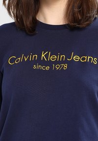Námořnická modrá bavlněná mikina s žlutým vyšitým nápisem: "Calvin Klein Jeans od 1978" na hrudi. Kulatý výstřih, ležérní střih.